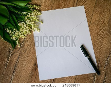 Bouquet Of Lilies Of The Valley On A Wooden Background And An Empty Piece Of White Paper With A Pen.