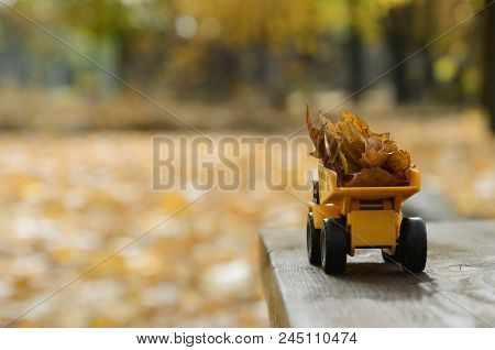 A Small Toy Yellow Truck Is Loaded With Yellow Fallen Leaves. The Car Stands On A Wooden Surface Aga