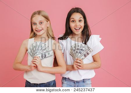 Photo of lucky caucasian girls 8-10 wearing casual clothing holding fans of dollars in hands isolated over pink background