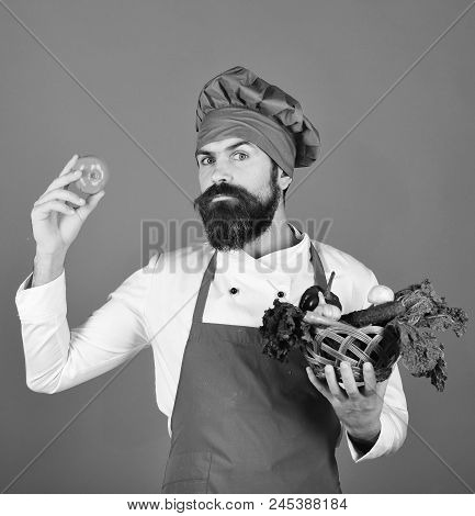 Mancook With Serious Face In Burgundy Uniform Holds Tomato And Vegetables In Wicker Bowl. Chef Holds