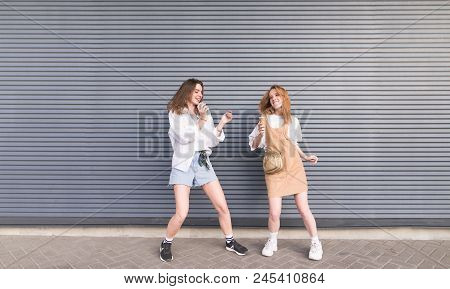 Two Young Girlfriends In Stylish Clothes Dance On A Gray Background. Portrait Of Two Cheerful Girls 