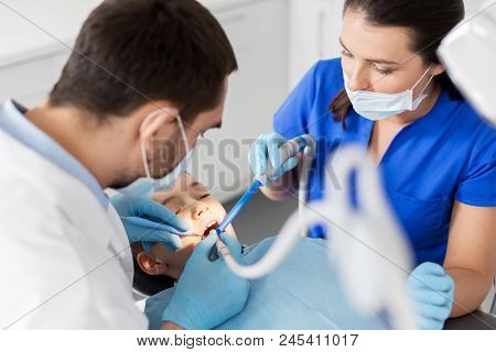 medicine, dentistry and healthcare concept - dentist and assistant with dental drill and saliva ejector treating kid patient teeth at dental clinic