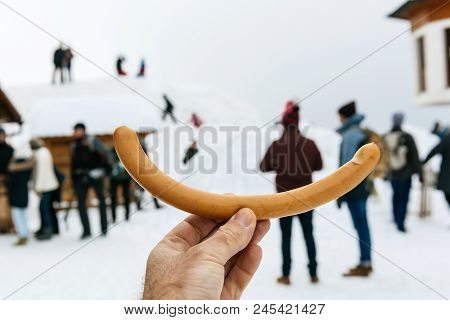 Male Hand Holding Sausage Point Of View At Long German Sausage With Silhouettes Of People Enjoying T