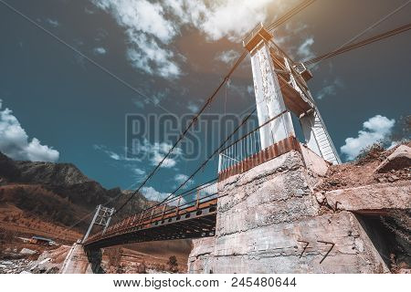 Close-up Wide-angle Shot Of The Suspension Bridge With Concrete Foundation Over The River In Mountai
