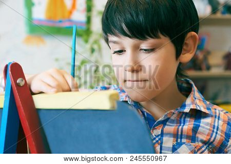 Child Standingnext To The Easel. Kid Boy Learn Paint By Brush In Class School. Kindergarten Interior