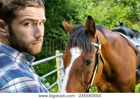 Good Looking, Hunky Cowboy Stands Next To Ranch With His Stallion, Horse And Saddle