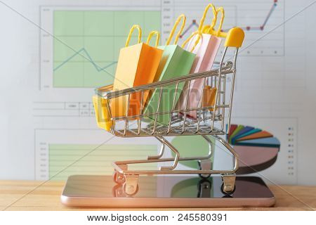 Colourful Paper Bags In Yellow Trolley On Smartphone With Chart Background On Table. Consumers Can B