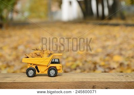 A Small Toy Yellow Truck Is Loaded With Yellow Fallen Leaves. The Car Stands On A Wooden Surface Aga