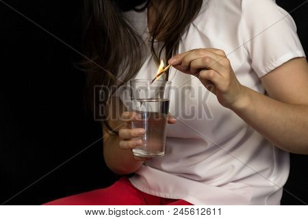 The Girl On A Black Background Holds A Glass Of Water And Extinguishes A Burning Match, Pain And Bur