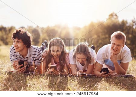 Cheerful Companions Smile While Tell Each Other Anecdotes, Use Modern Smart Phones, Have Picninc Out