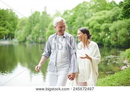 Happy seniors taking walk along waterside and talking about some pleasant things