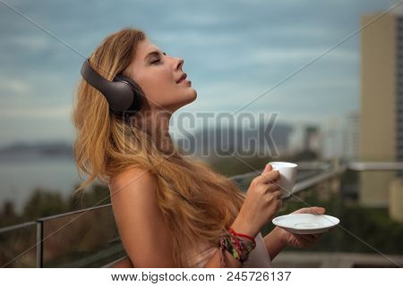 Young girl drinking coffee and listening music with headphones on the terrace