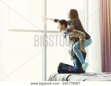 Loving Young Couple Looking Through Window In Hotel Room. Happy Woman Pointing Away