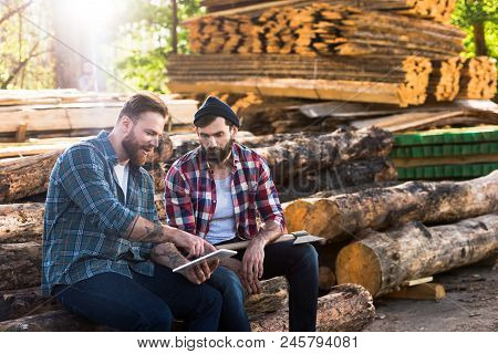 Bearded Lumberjacks Sitting On Logs With Axe And Digital Tablet At Sawmill