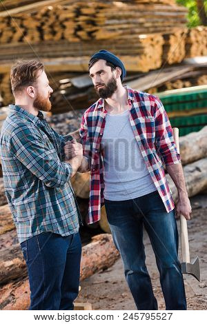Bearded Lumberjack In Checkered Shirt Holding Axe And Shaking Hands With Partner At Sawmill
