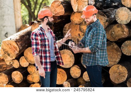 Lumberjack In Protective Helmet Pointing By Finger On Logs To Partner While He Writing In Clipboard