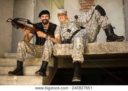 The Smiling Male And Female Army Soldiers Are Posing In Military Uniform With Crossbow Weapon Indoor