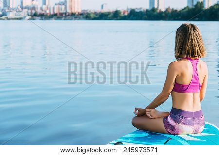 Young Woman Meditating On Paddle Board. Practice Yoga On Paddle Board, Woman Sitting On Paddle Board