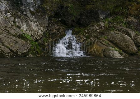 The Spring Follows A Waterfall From The Rock And Flows Into The River On A Precipitous River Bank