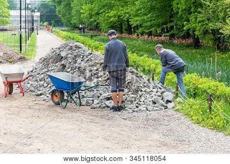 Two Workers Load Stones Into A Wheelbarrow During The Repair Of The Sidewalk In The Park