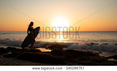 Surfer Girl Holding Surfboard On The Beach. Silhouette Of Surfer Girl During Sunset Time. Golden Hou