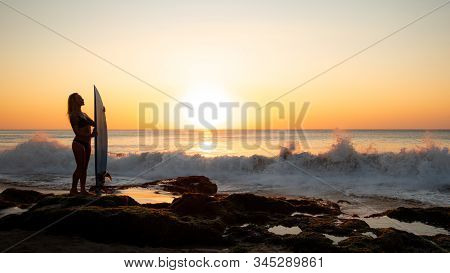 Surfer Girl Holding Surfboard On The Beach. Silhouette Of Surfer Girl During Sunset Time. Golden Hou