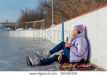 Lovely Young Woman Relaxing After Riding Ice Skates And Drinking Hot Drink From Termo Pot On The Ice