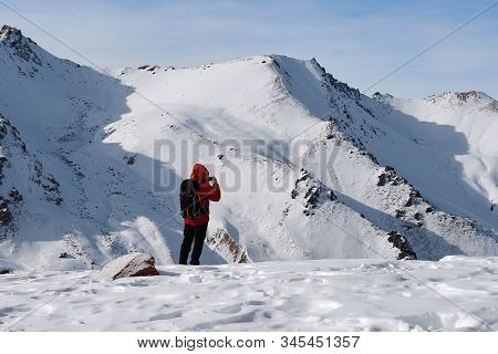 Man In Red Jacket With Black Backpack Is Standing At Mountain Plateau Against The High Snowy Mountai