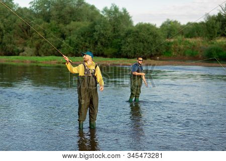 Two Fishermen Are Standing In The River, Wearing Rubber Boots. Men Throw Fishing Rods. Fishing Adven