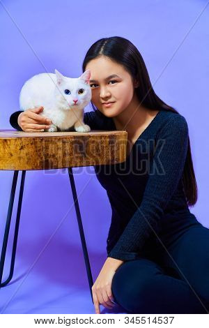 A Girl Of Asian Appearance Sits On The Floor Next To A Wooden Table - On The Table Sits A White Cat 
