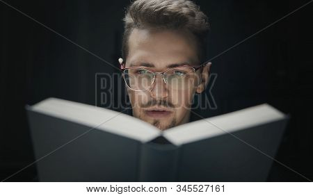 Close-up Of Serious Young Bearded Man In Glasses Behind Book Isolated On Black Background