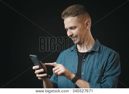 Smiling Young Man In Jean Shirt Holding Smartphone With One Hand And Touching Screen With The Other,