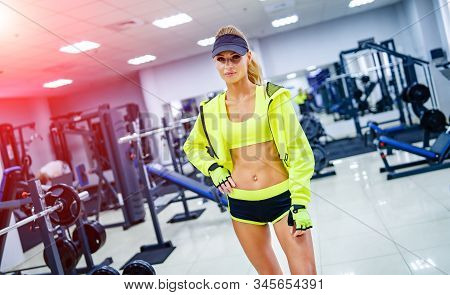 Gorgeous Blonde Woman Is Standing At A Gym Looking At Camera. Wearing Bright Sports Wear And A Cap. 
