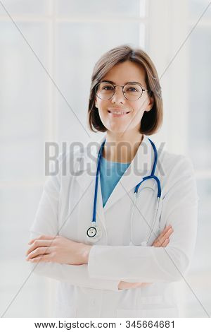 Beautiful Cheerful Woman Doctor Wears White Gown, Glasses And Phonendoscope, Keeps Hands Crossed, Lo