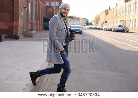 Handsome Young Man In Grey Coat And Hat Crossing The Street With A Cup Of Coffee.