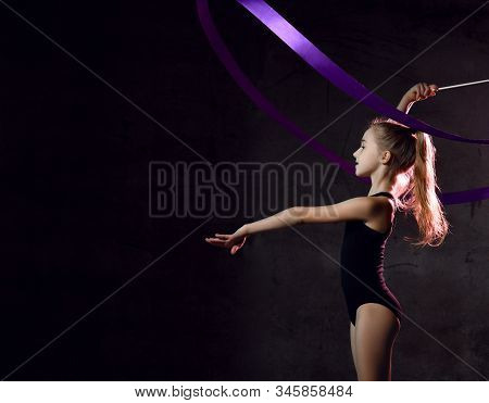 Young Girl Gymnast In Black Sport Body And Uppers Standing Sideways And Making Exercise With Blue Gy