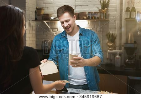 Barista Man Standing Behind The Bar Sells Hot Coffee Drink In Paper Cup To Woman Client For Takeaway