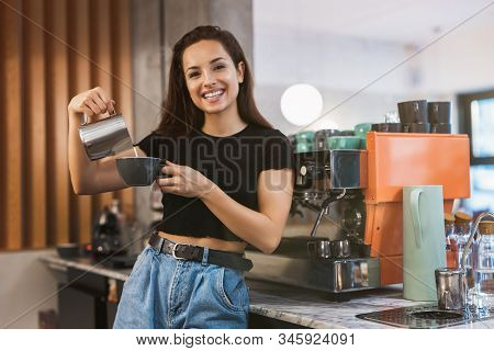 Young Smiling Barista Womanstanding Behind The Bar Pouring Whipped Milk Into Coffee Drink, Preparing