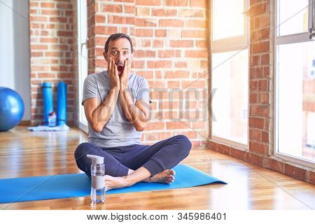 Middle age handsome sportman sitting on mat doing stretching yoga exercise at gym afraid and shocked, surprise and amazed expression with hands on face