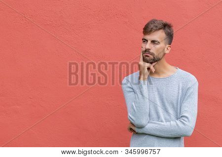 Young Bearded Man In Thoughtful Pose With Fingers On Chin On Red Wall Background With Copy Space. Mi