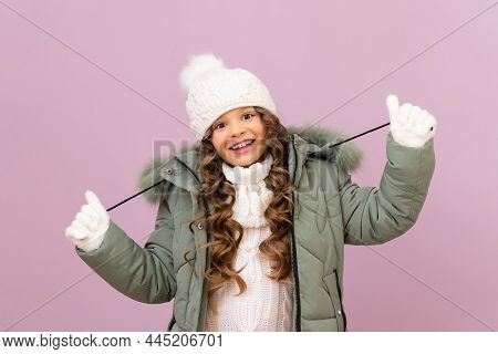 A Little Girl In A Warm Jacket And A Knitted White Hat With Curly Hair. A Child In A Warm Winter Jac