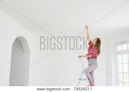 Full-length of woman on ladder fitting light bulb in new house