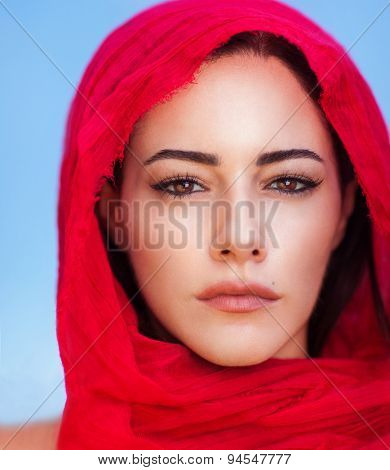 Closeup portrait of beautiful arabic woman wearing red headscarf over blue sky background, perfect natural makeup, traditional arabian beauty