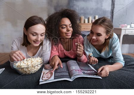 What do you think about this. Cheerful smiling involved girls reading the magazine while lying on the bed at home and eating popcorn