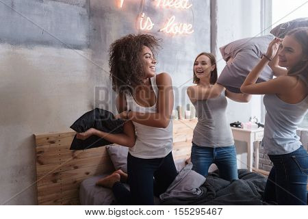 We playing funny games. Amused laughing young girls standing on the bed in the bedroom while having a funny pillow fight and expressing joy
