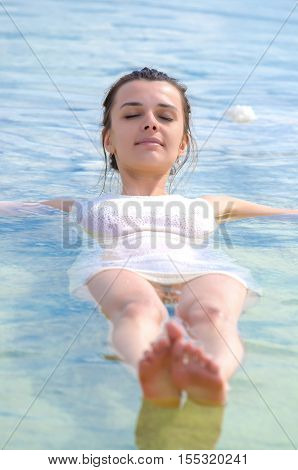 Sexy attractive girl wears white dress swims in water and looks to camera. Her hands and legs lowered into emerald sea. Young woman relaxing sitting on the edge of Baskunchak salt lake on white crystals of salt. Fron to camera