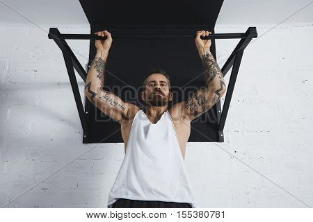 Strong tattooed in white unlabeled tank t-shirt male athlete shows calisthenic moves close up Pullup hold on medium move on pull bar