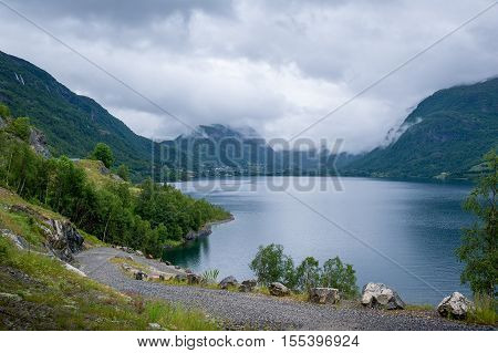 Gravel serpentine road at scenic norwegian fjord shore. Mountains and clouds - typical Norway landscape.