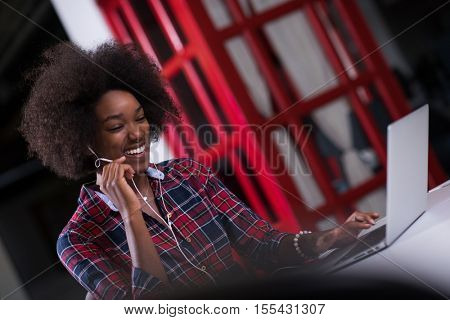 black woman in modern office at her workplace speeking on phone over earphones and working on laptop computer
