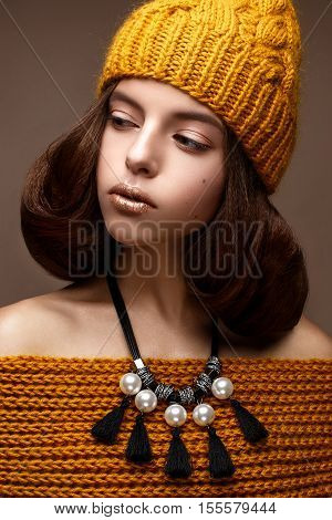 Beautiful girl in a knitted hat on her head and a necklace of pearls around her neck. The model with gentle make-up and gold lips. Beautiful face. Photo taken in a studio. Gentle and cozy winter look.